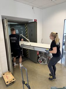 Zwei Personen tragen ein großes weißes Möbelteil zu einem geöffneten Aufzug