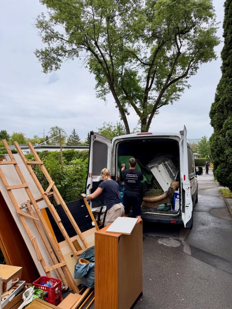 Zwei Personen in Firmenkleidung verladen Möbelplatten und einen Lattenrost in einen weißen Transporter